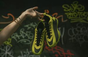A hand holding up a pair of A.E. 1 Low 'Preloved Lime' sneakers in vibrant lime green with black accents, against a graffiti-covered urban background